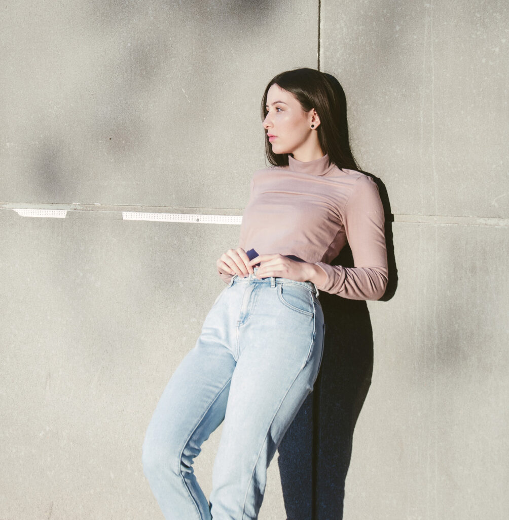 Gabriela Inzunza stands against a gray concrete wall leaning slightly to her right with her long brown hair cascading down her back. She wears a muted pink long-sleeve top and light blue high-waisted jeans paired with black ankle boots.
